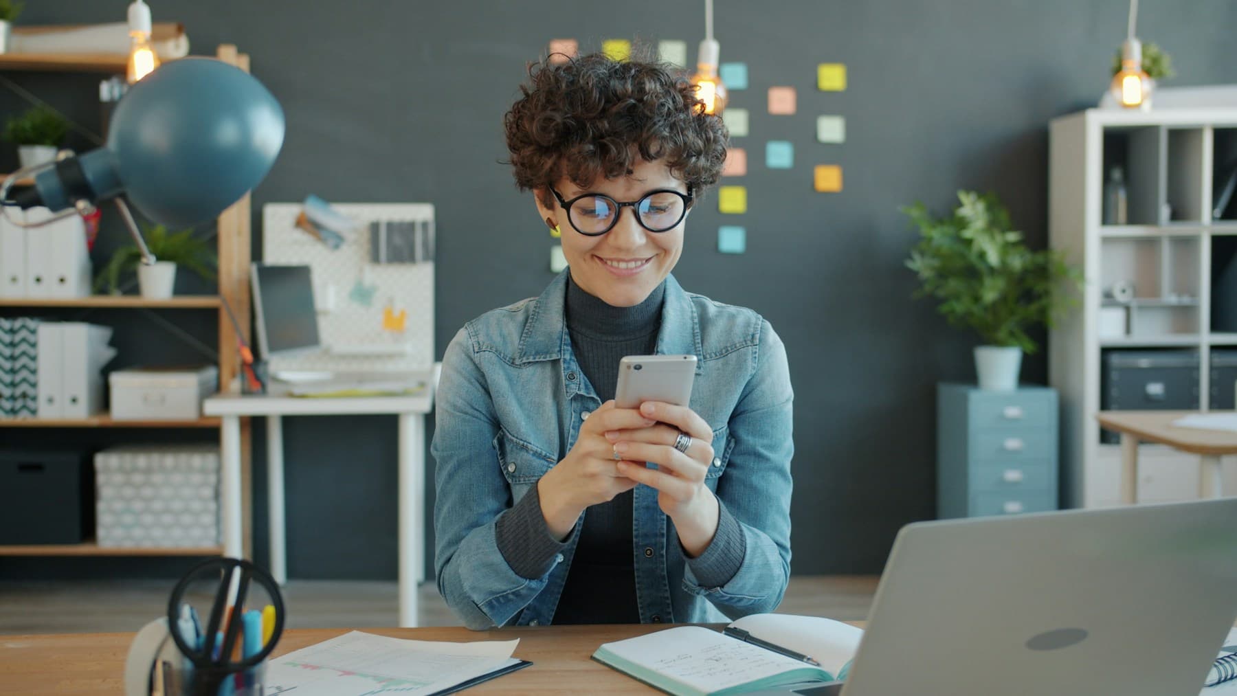 A woman in glasses checking her phone at her desk